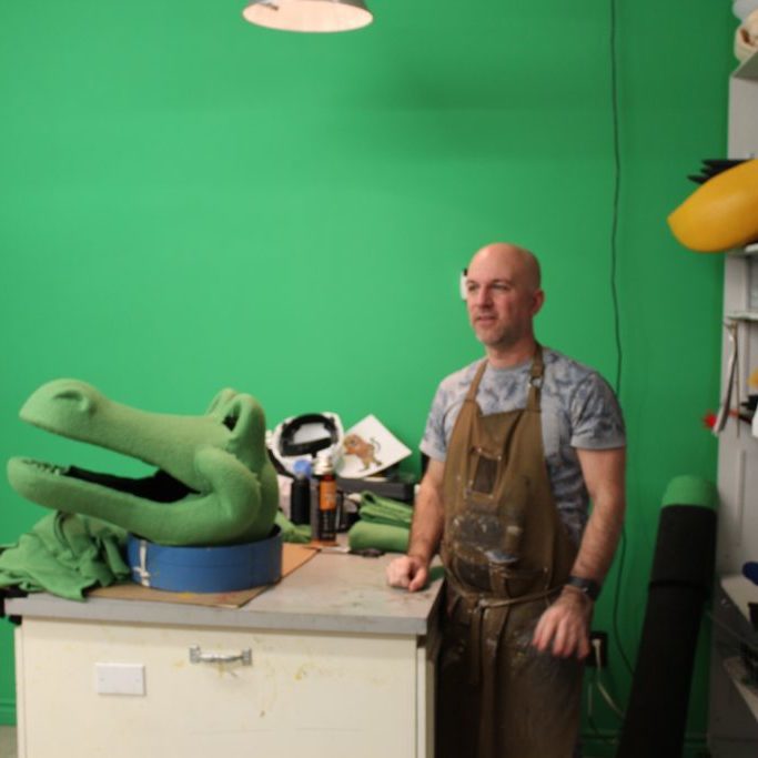 Man in a grey t-shirt and a leather apron next to a mascot crocodile head in a green room.
