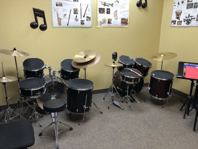 Two full drum kits sitting against a yellow wall. There are posters that discuss different instruments above the drums.