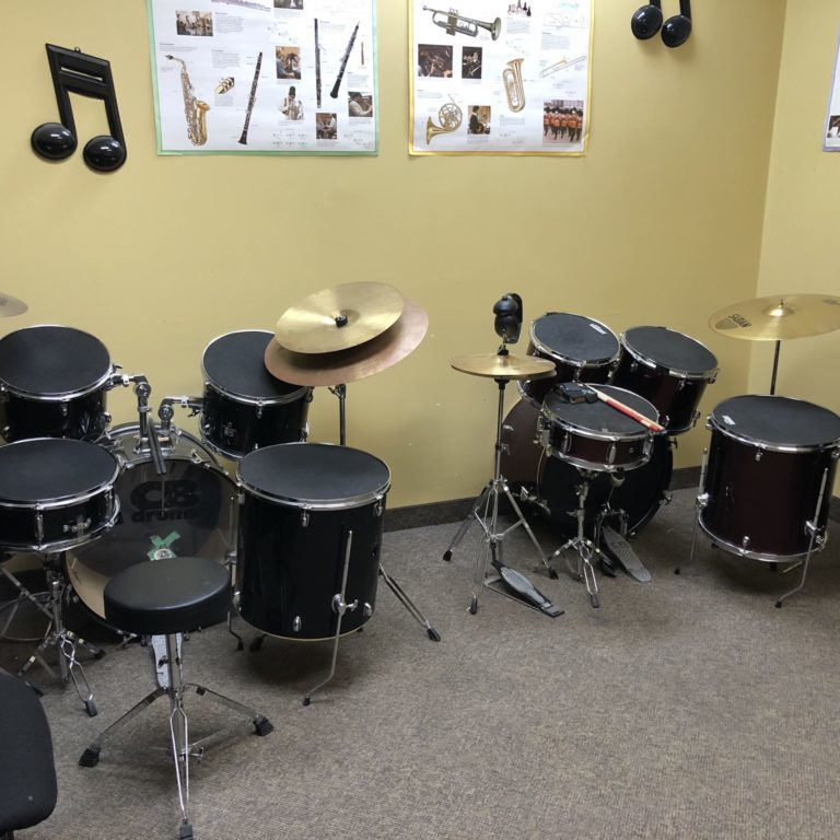 Two full drum kits sitting against a yellow wall. There are posters that discuss different instruments above the drums.