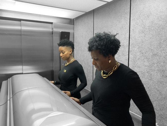 Two women are standing in front of a silver casket in an elevator. They are both in gold jewelry and black shirts.