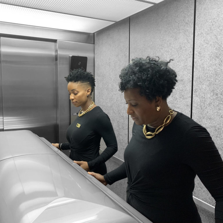Two women are standing in front of a silver casket in an elevator. They are both in gold jewelry and black shirts.