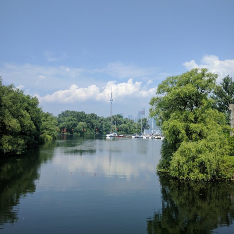 Far away photo of the CN tower surrounded by trees and water with clouds in the background at a harbour during the daytime