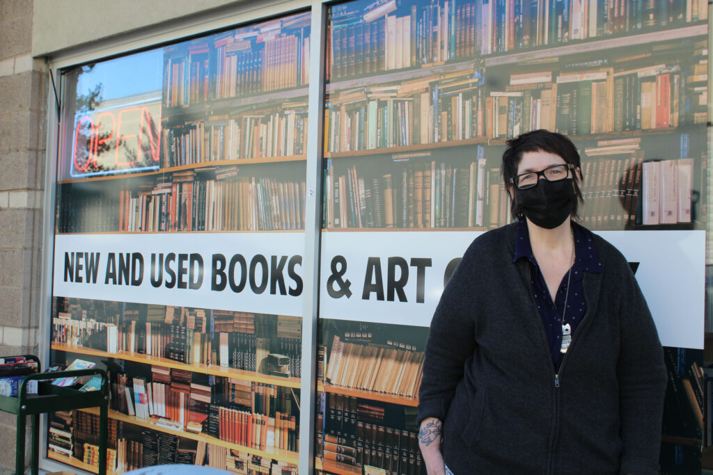 Woman stands outside with sign reading "new and used books and art" behind her.