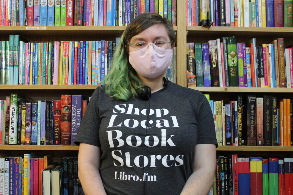 A woman wearing a "shop local book stores" shirt stands in front of a full bookshelf.