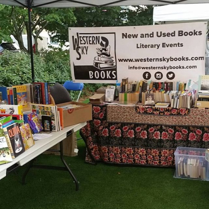 Tables filled with books sit on green grass with a banner for Western Sky Books hanging from the top of a tent.