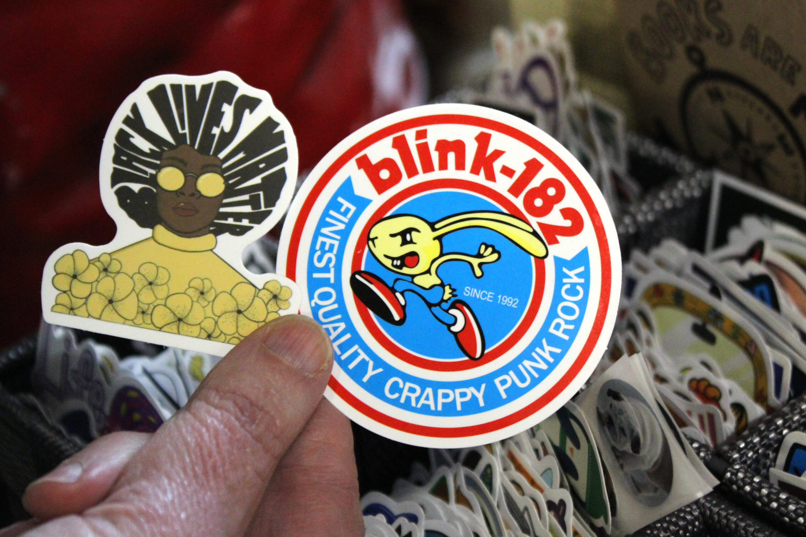 A hand holds two stickers: one with a Black Lives Matter design and the other a Blink-182 design.