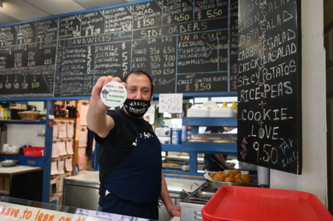 Man in a mask holding a button pin that says "Mystic Muffin".