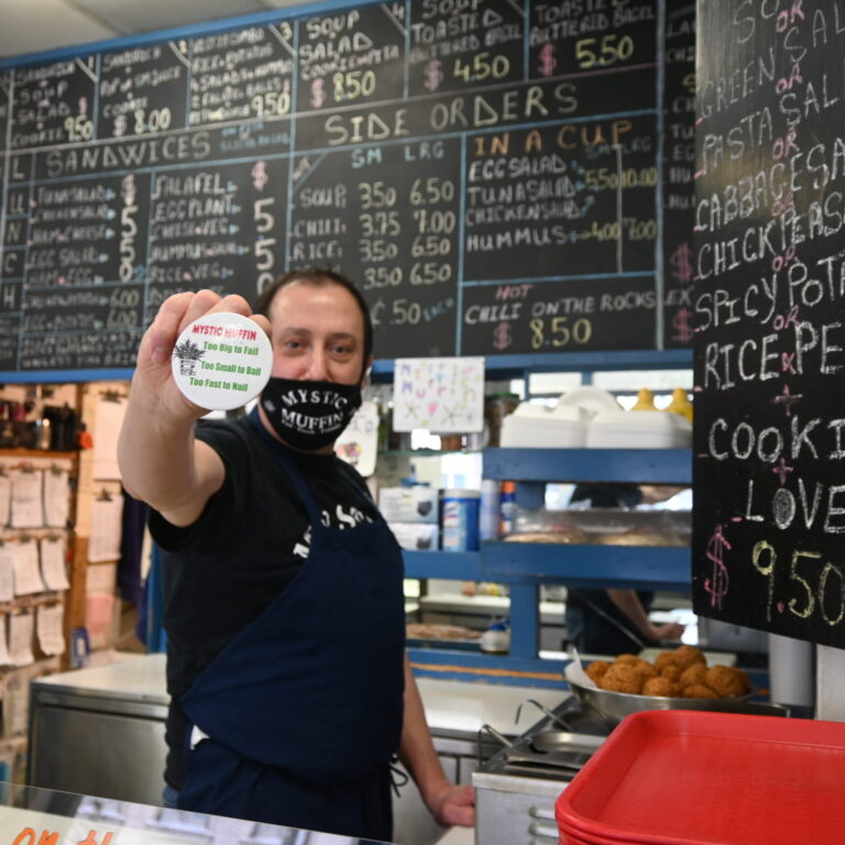 Man in a mask holding a button pin that says "Mystic Muffin".