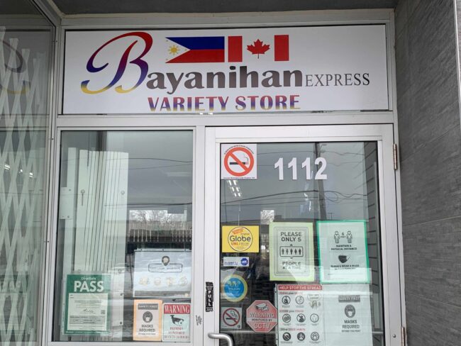 The metal door entrance to Bayanihan Express. On the top is their store banner. There are multiple posters on the glass of the metal-framed door.