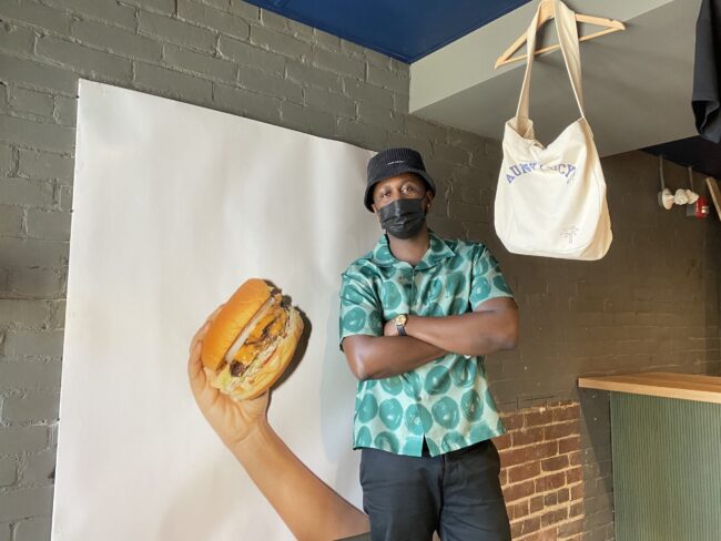 A man wearing a mask, teal-patterned shirt and a black bucket hat standing with his arms crossed in front of a poster with a hand holding a burger on a white background with a tote bag hanging on the man's left-hand side.