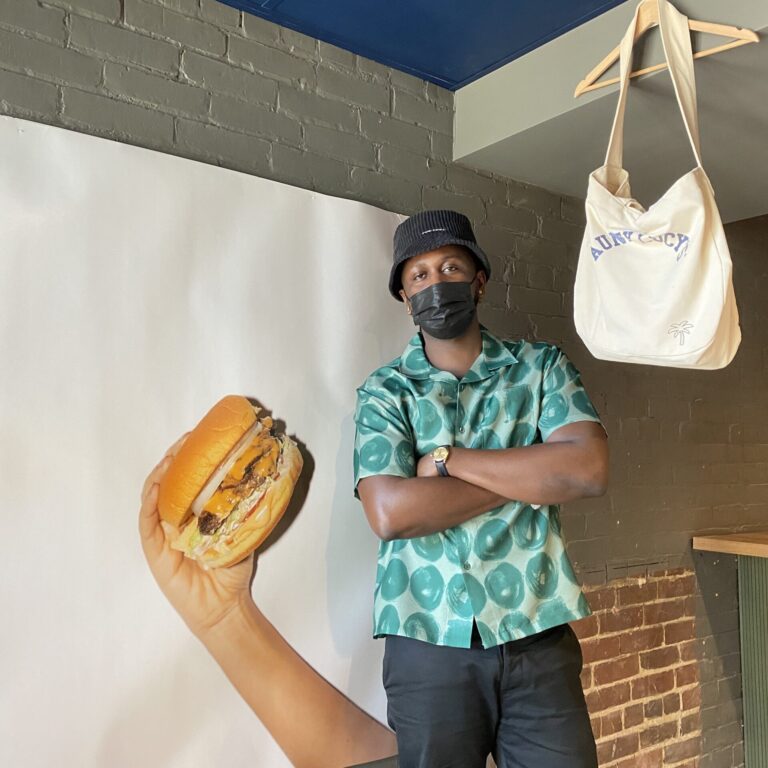 A man wearing a mask, teal-patterned shirt and a black bucket hat standing with his arms crossed in front of a poster with a hand holding a burger on a white background with a tote bag hanging on the man's left-hand side.