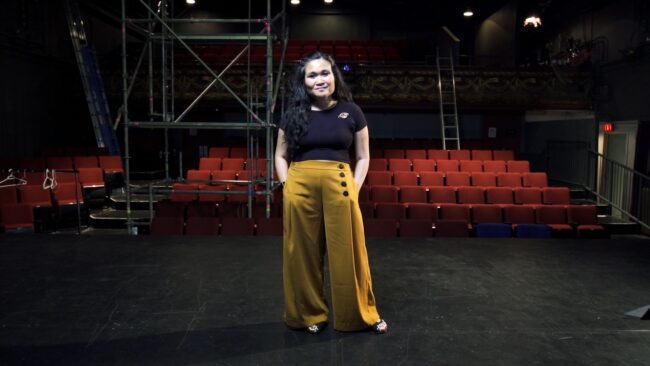 Woman in a black t-shirt and yellow pants standing in an empty theatre.