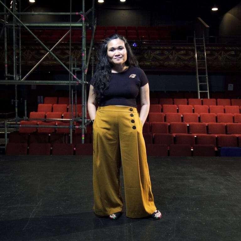 Woman in a black t-shirt and yellow pants standing in an empty theatre.