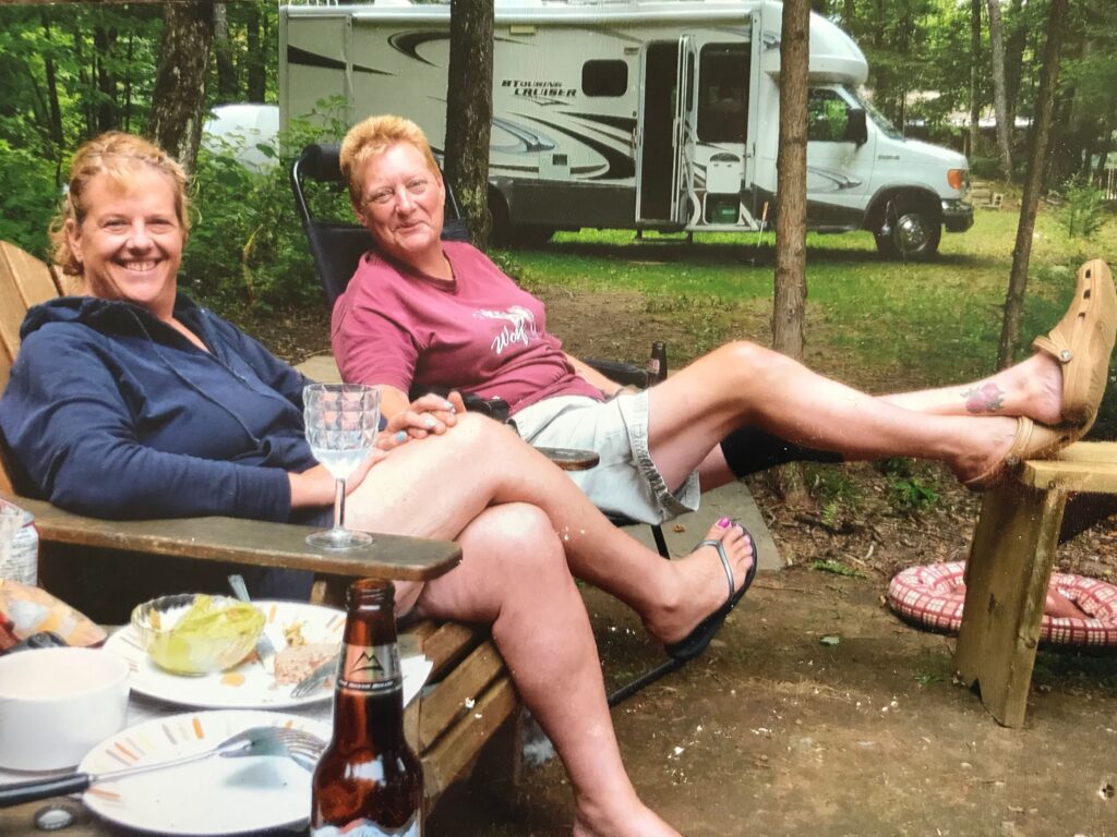 Lori Pilatzke and Jen Maklin sit in Muskoka chairs on a campsite, their RV visible in the background.