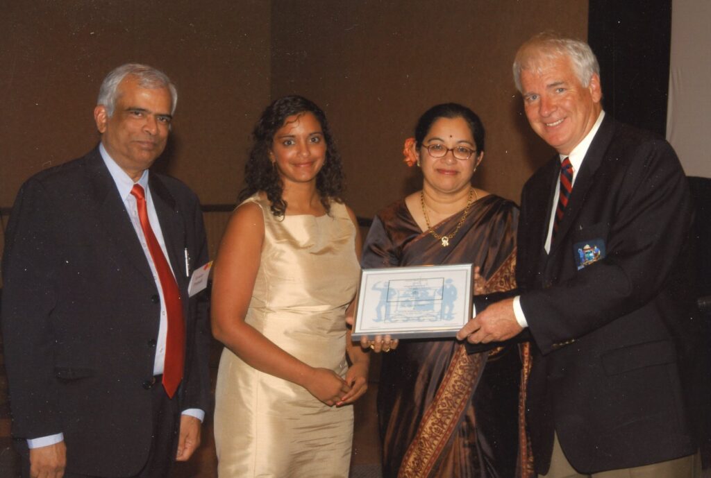 Eswar and the mayor wear black suits and Niru and Shashi wear gold and brown dresses as the mayor holds an award.
