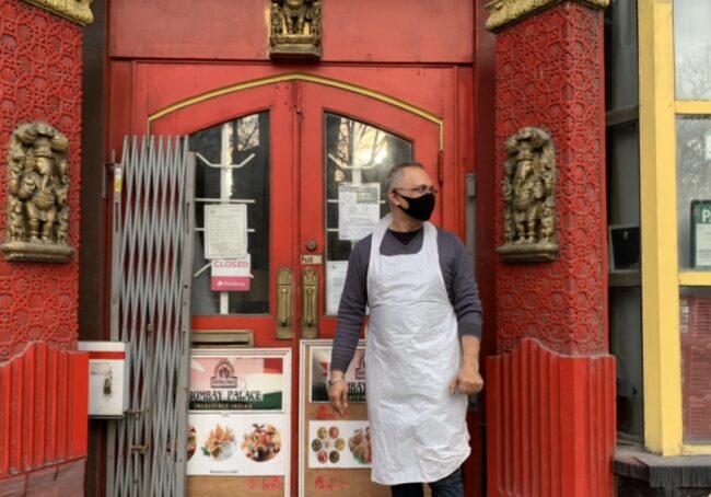 Man in an apron in front of a restaurant door.