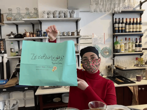 A staff member smiling at the camera while carrying a blue paper bag which says, "Zezafoun Syrian Cuisine" followed by the phone number and email of the restaurant. 