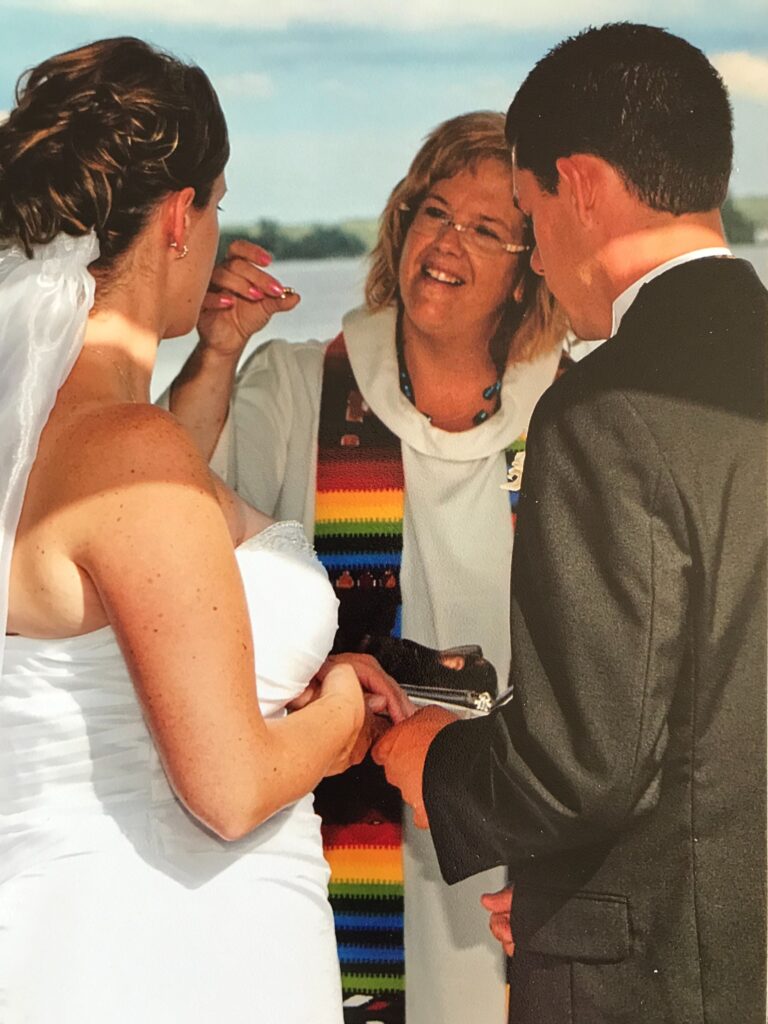 Lori Pilatzke performs a wedding, smiling and holding a wedding ring, as the couple stands in front of her.