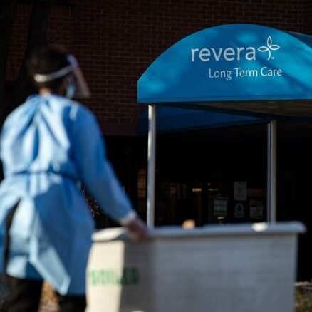 Woman in PPE and a face shield wheeling a container to a building.