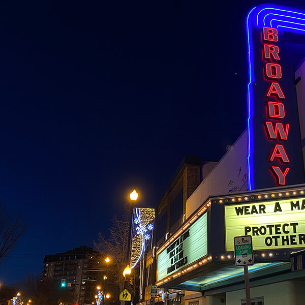 A city at night. Neon sign with the words "Broadway", and then a sign that says "Wear a Mask. Protect Each Other" under it.