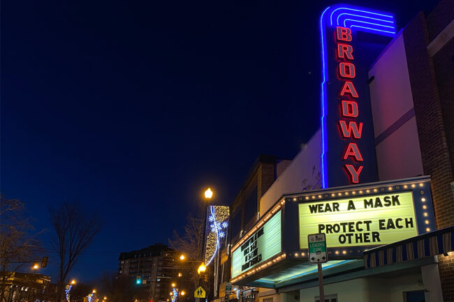 A city at night. Neon sign with the words "Broadway", and then a sign that says "Wear a Mask. Protect Each Other" under it.