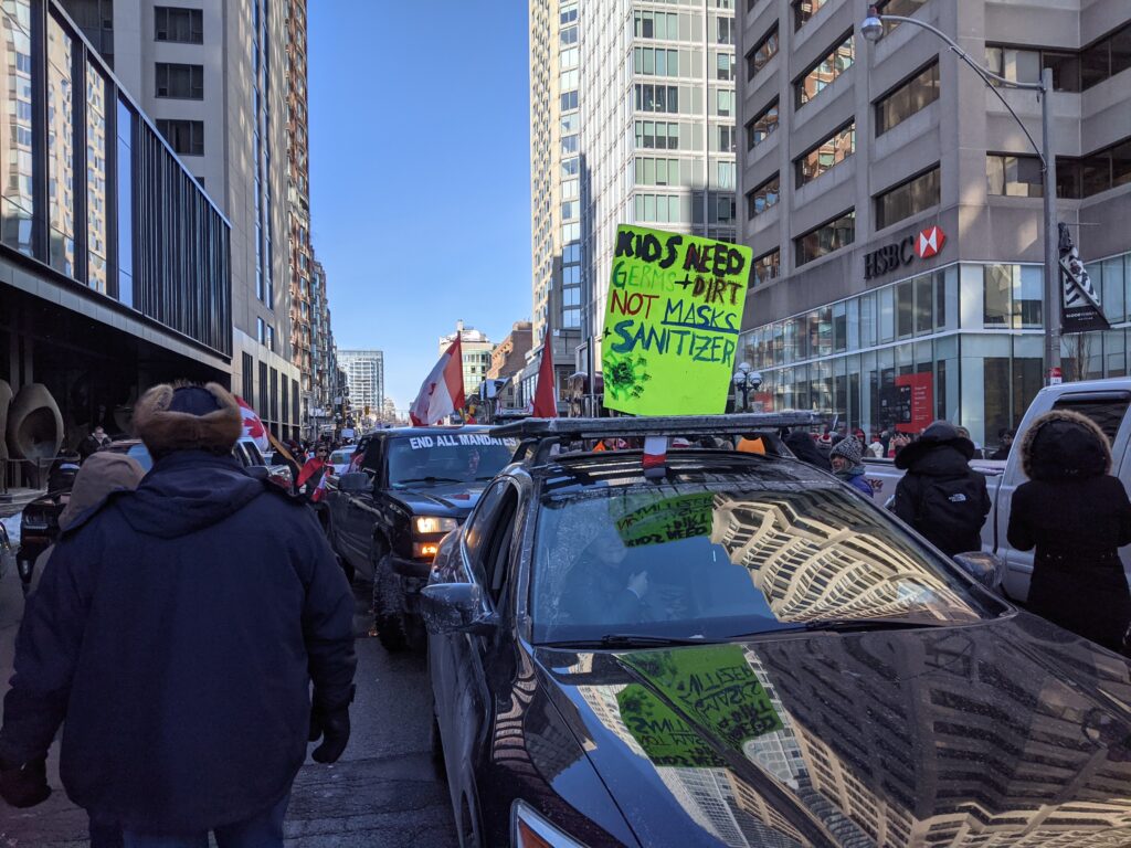 Rows of cars and trucks drive through a city street with people walking alongside them, raising flags and posters with protest messages written on them.