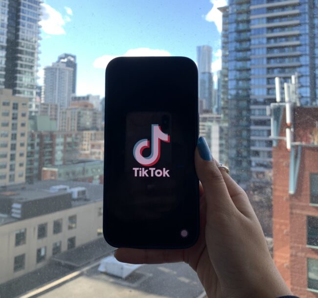 A hand holding a smartphone showing the TikTok logo in front of a window