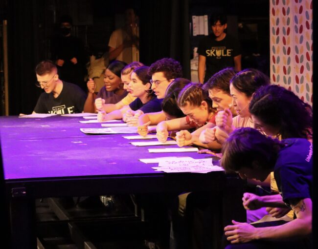 Students sitting at a table under colourful lights looking confused and stressed.