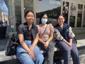 3 students sitting on a bench outside.  Two are maskless and one in the middle is wearing a blue mask.