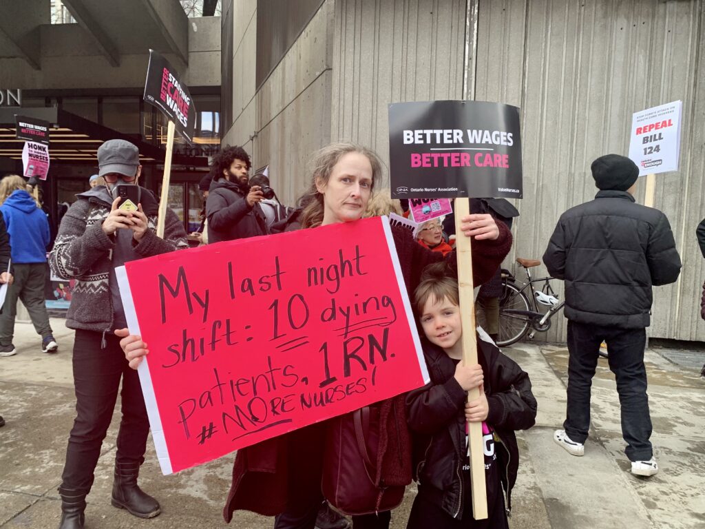 A woman and a young boy hold up two signs. One is a neon pink sign that says, “My last night shift: 10 dying patients. 1 RN. #MoreNurses!” The other one is black and says, “Better wages, better care.”