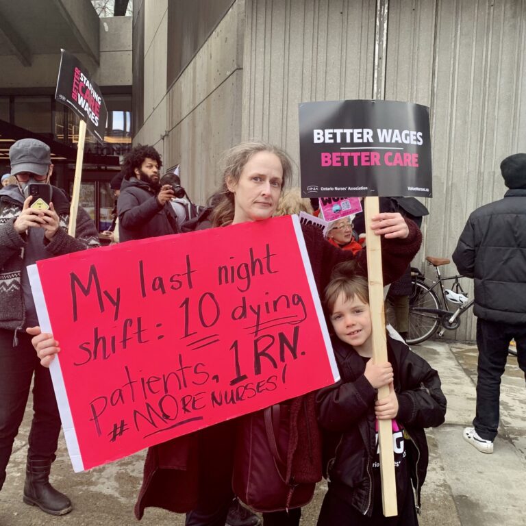 A woman and a young boy hold up two signs. One is a neon pink sign that says, “My last night shift: 10 dying patients. 1 RN. #MoreNurses!” The other one is black and says, “Better wages, better care.”