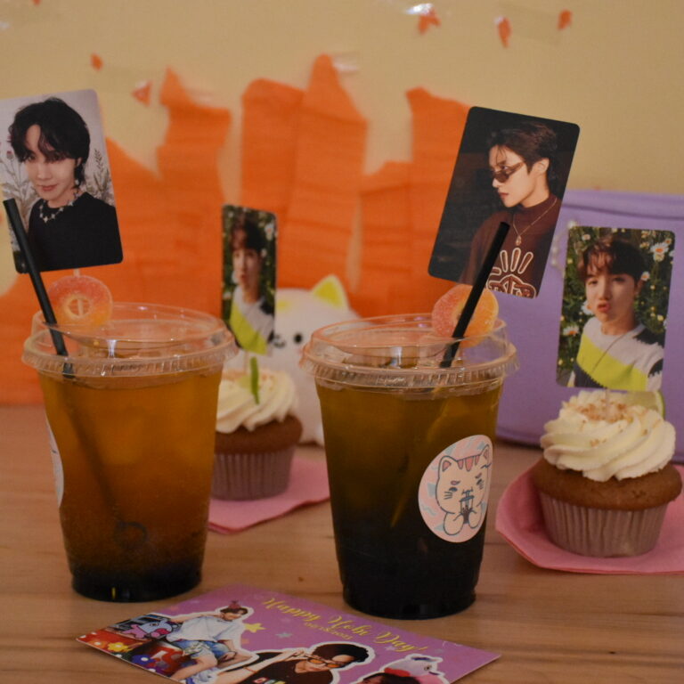 Two cups filled with juice on top of a wooden table with photographs of Jung Hoseok from BTS displayed along decorative birthday-themed postcards