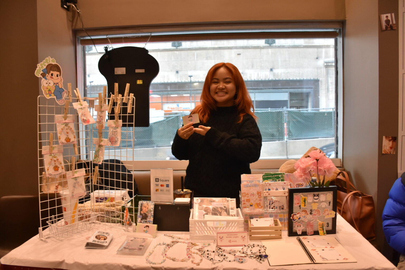 Girl with red hair poses in front of K-pop-themed stationary products and handmade keychains at a local bubble tea shop