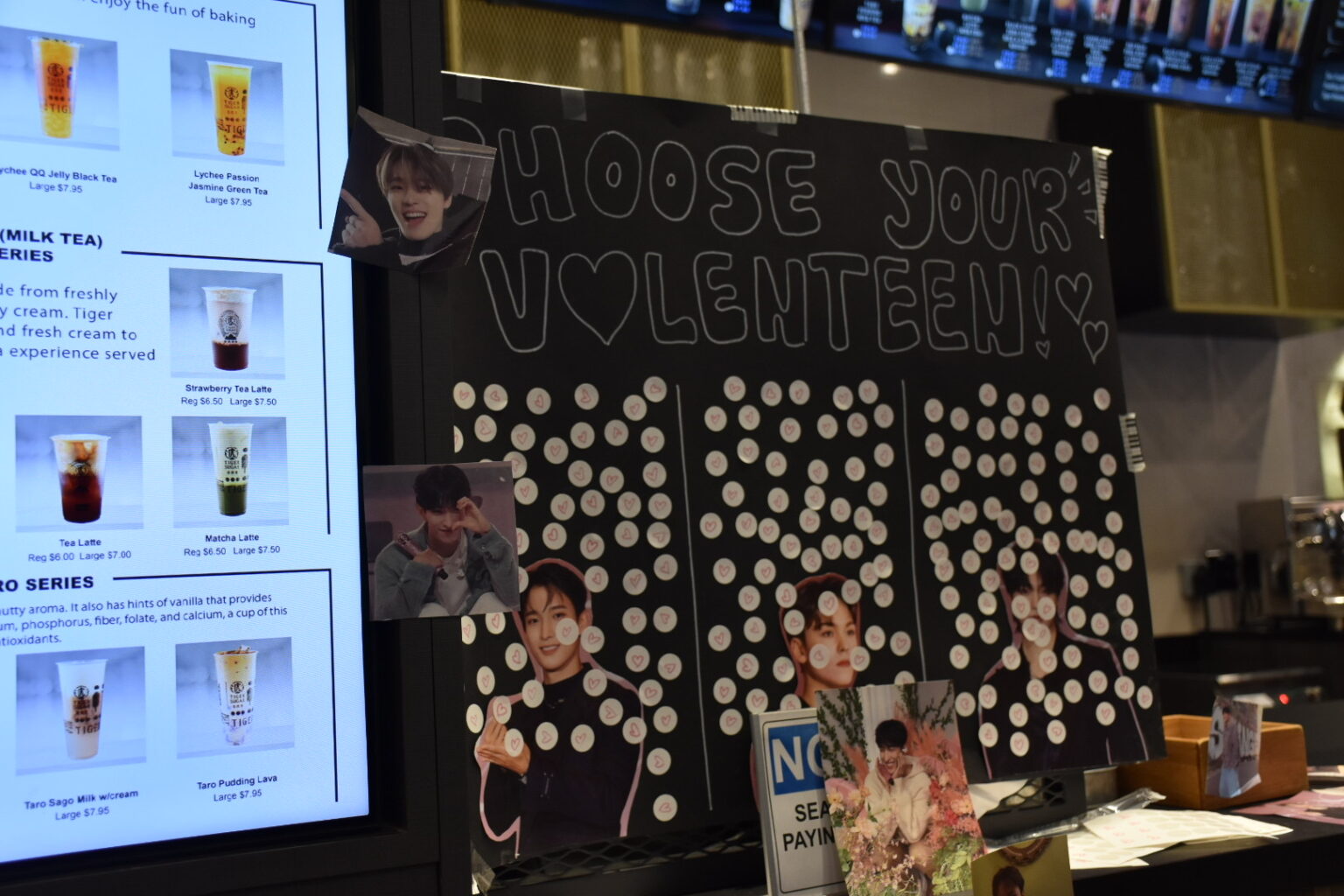 A black posterboard titled “Choose Your Valenteen” sits on the counter of a local bubble tea cafe with white stickers on K-pop group members from SEVENTEEN decorated by fans