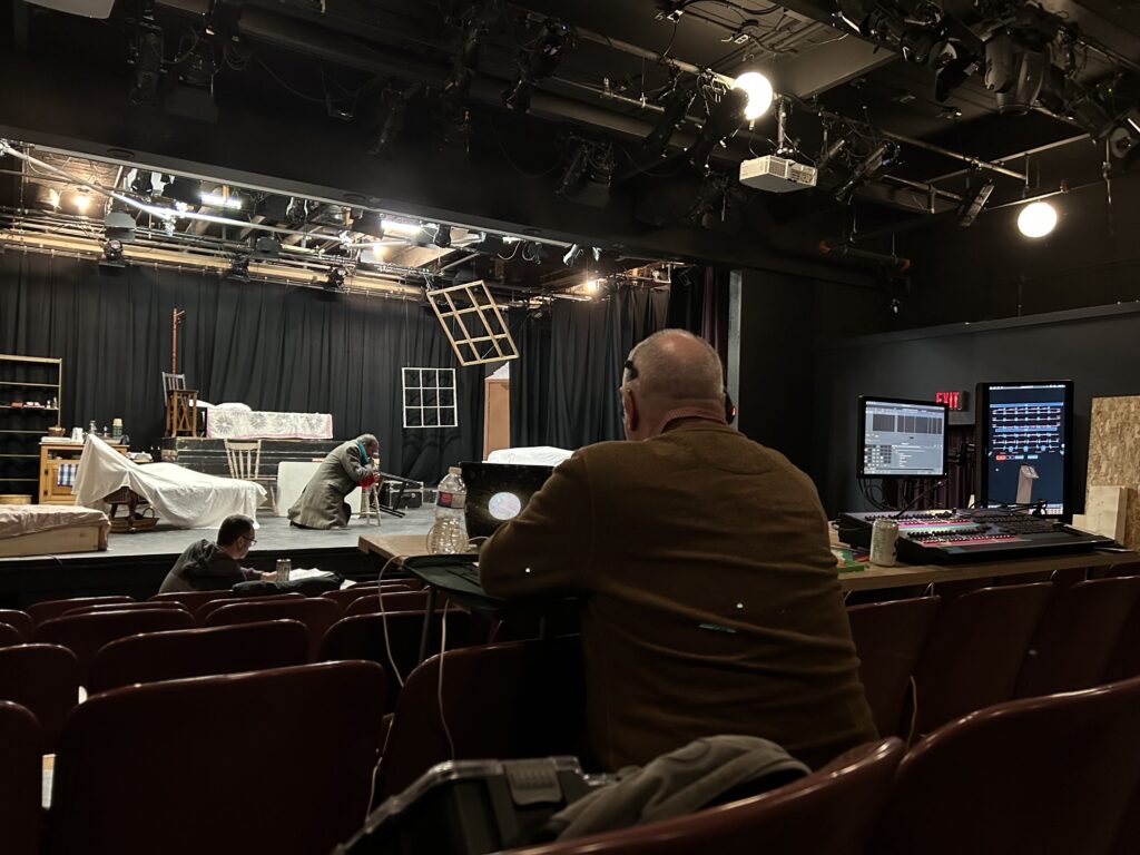 A man sits behind a desk, working on a computer while sitting on maroon-coloured theatre seats in a nearly empty auditorium