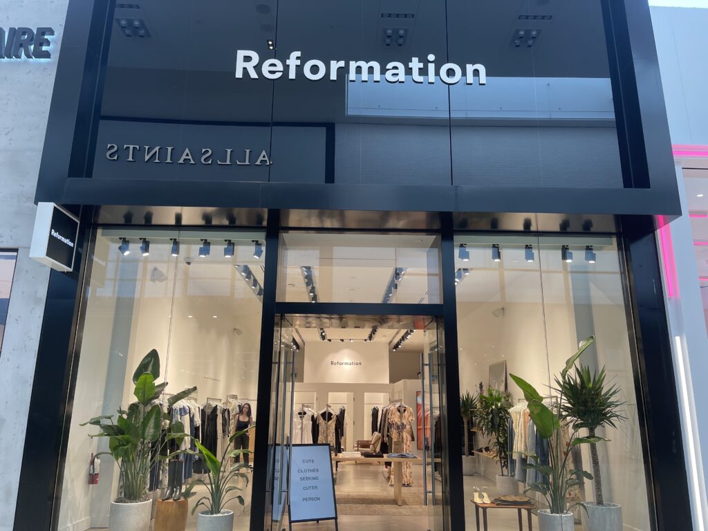 Front entrance of Reformation at Yorkdale Shopping Centre. There are plants in the window and a sign in the distance that reads, "Cute Clothes Seeking Cuter Person."