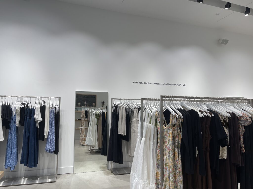 Overview of the whole Reformation store (including a mirror and racks of clothing) with their slogan that reads, "being naked is the #1 most sustainable option. We're #2."