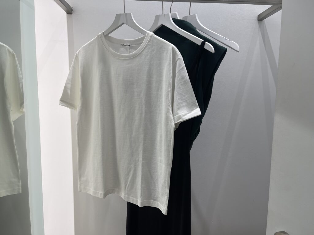 A white t shirt on a hanger in a fitting room with clothing items behind it.