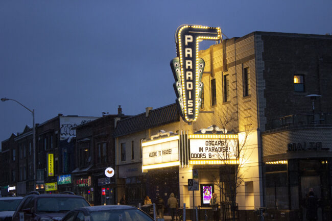 The exterior of the Paradise Theatre cinema at night. Bright lightbulbs line the marquee, which says "Welcome to Paradise!" and "Oscars in Paradise! Doors 6PM." A few people walk past the theatre, and one stands calmly outside the front door.