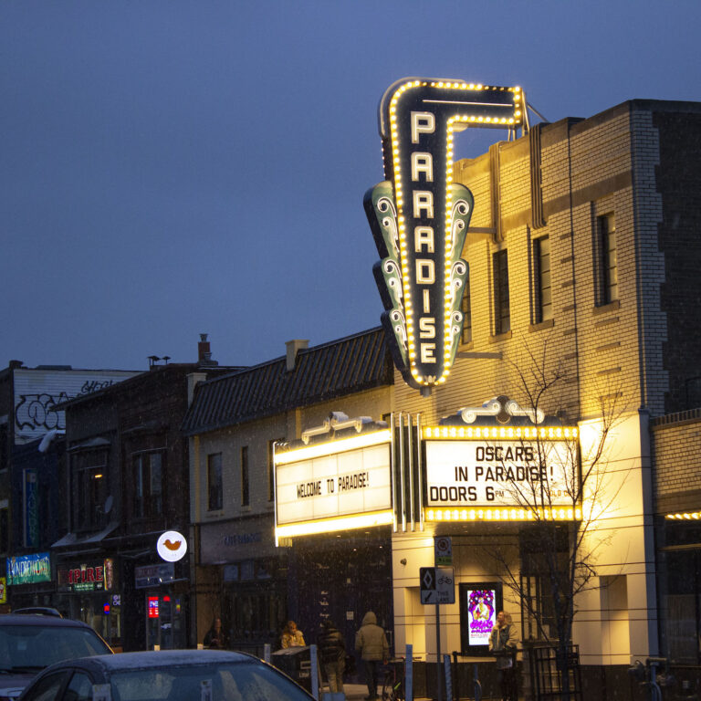The exterior of the Paradise Theatre cinema at night. Bright lightbulbs line the marquee, which says "Welcome to Paradise!" and "Oscars in Paradise! Doors 6PM." A few people walk past the theatre, and one stands calmly outside the front door.