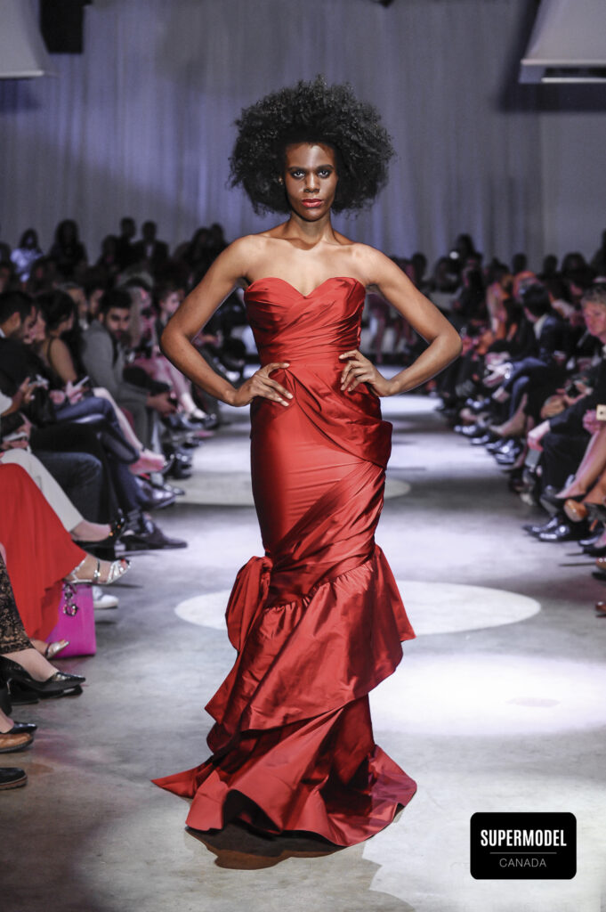 Nini Amerlise wearing a long red off the shoulder dress on a runway