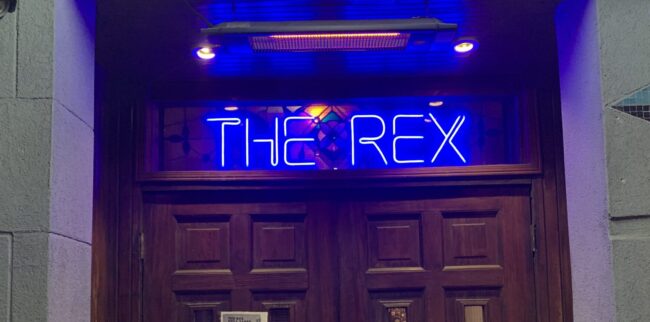 A neon sign above a wooden outside door reads "The Rex"