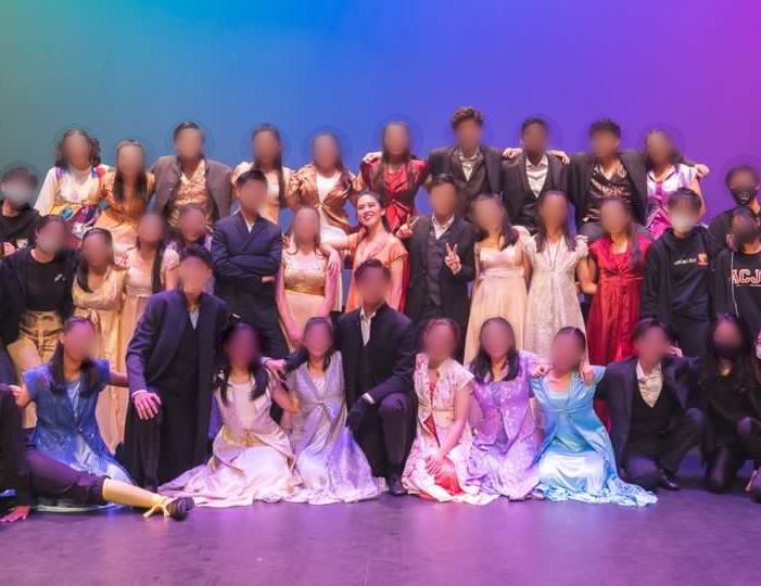 A large group of actors in costume pose together on a stage. Micah tan is in the middl eof the group.