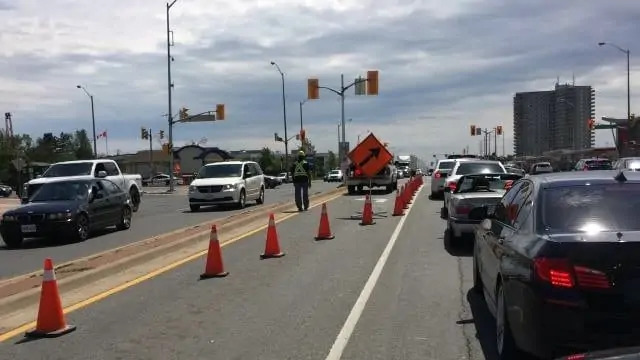 Orange traffic cones blocking cars on the road with a worker in a bright safety vest and helmet.