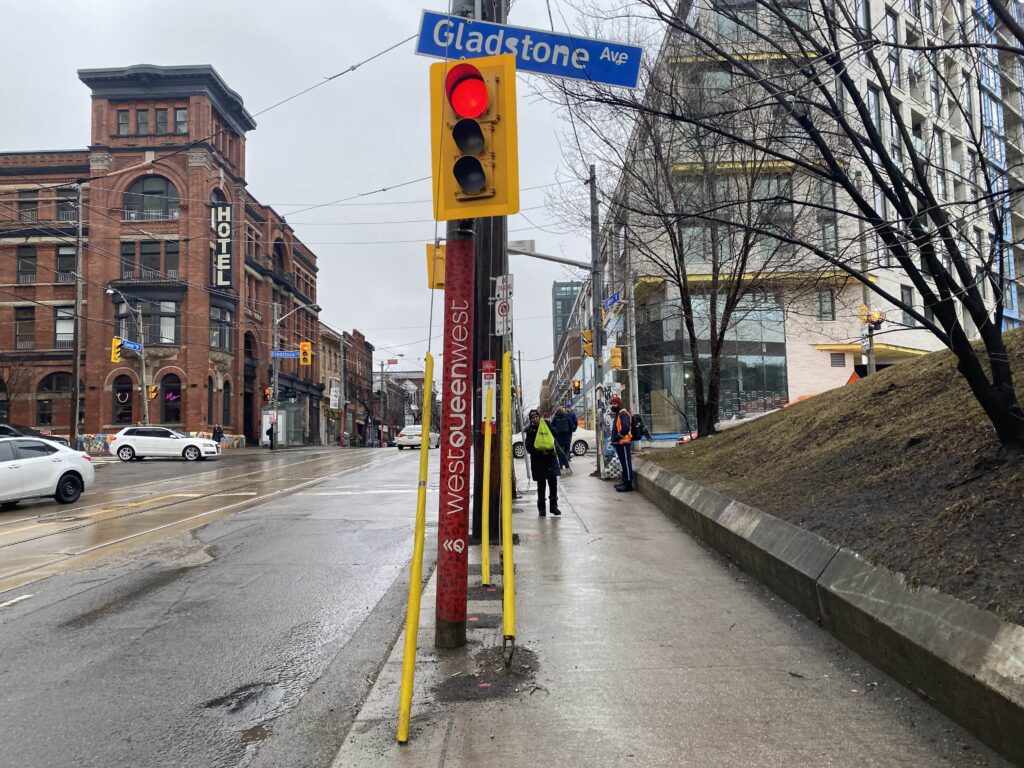 An picture of a Queen Street West sign next to a red light on a traffic sign on a rainy day with wet roads.