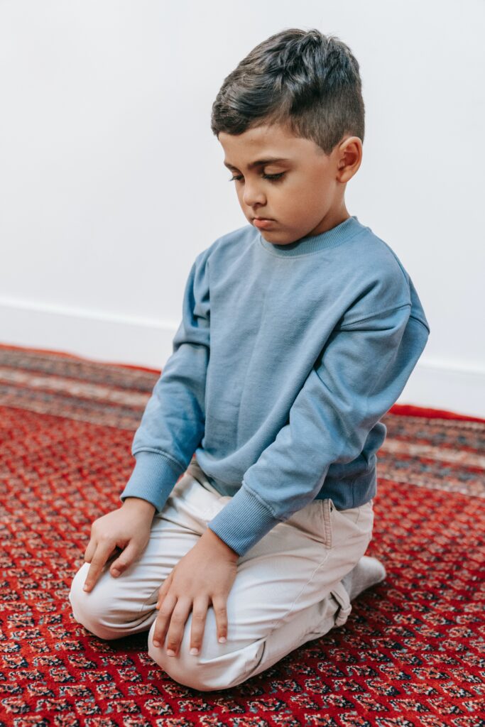 Child in blue sweater kneels on the floor in prayer.