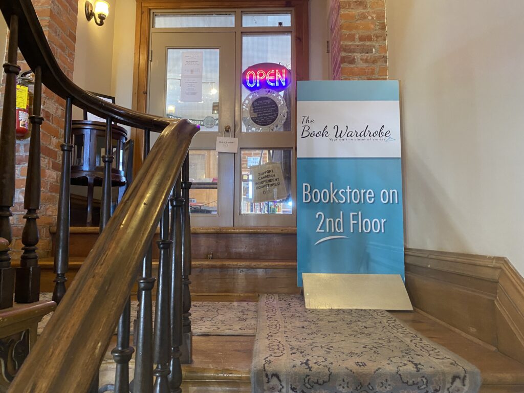 A gray door sits atop a number of carpeted steps. In front of the door, a blue sign advertises The Book Wardrobe.