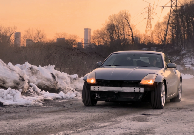 Image of a silver Nissan 350Z parked on a dirt trail at early sunset. The car is missing its front bumper, revealing the engine and underside. The surrounding landscape includes trees and residential complexes in the distance.