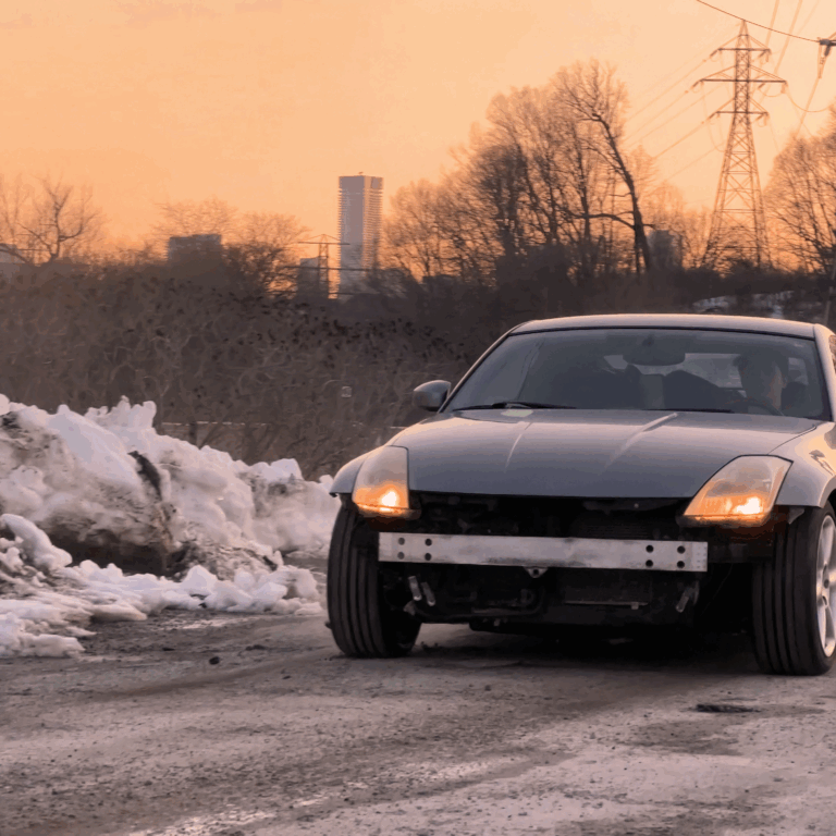 Image of a silver Nissan 350Z parked on a dirt trail at early sunset. The car is missing its front bumper, revealing the engine and underside. The surrounding landscape includes trees and residential complexes in the distance.