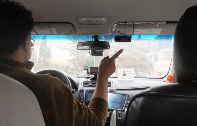 Photo from the backseat of a taxi featuring the driver pointing out the window.